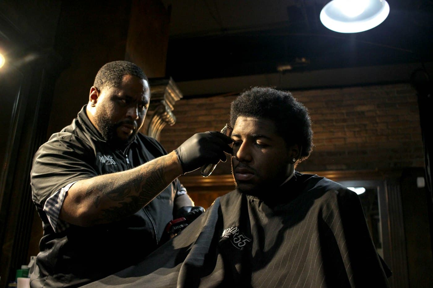 A barber in gloves lines up a client's hairline in a warmly lit shop