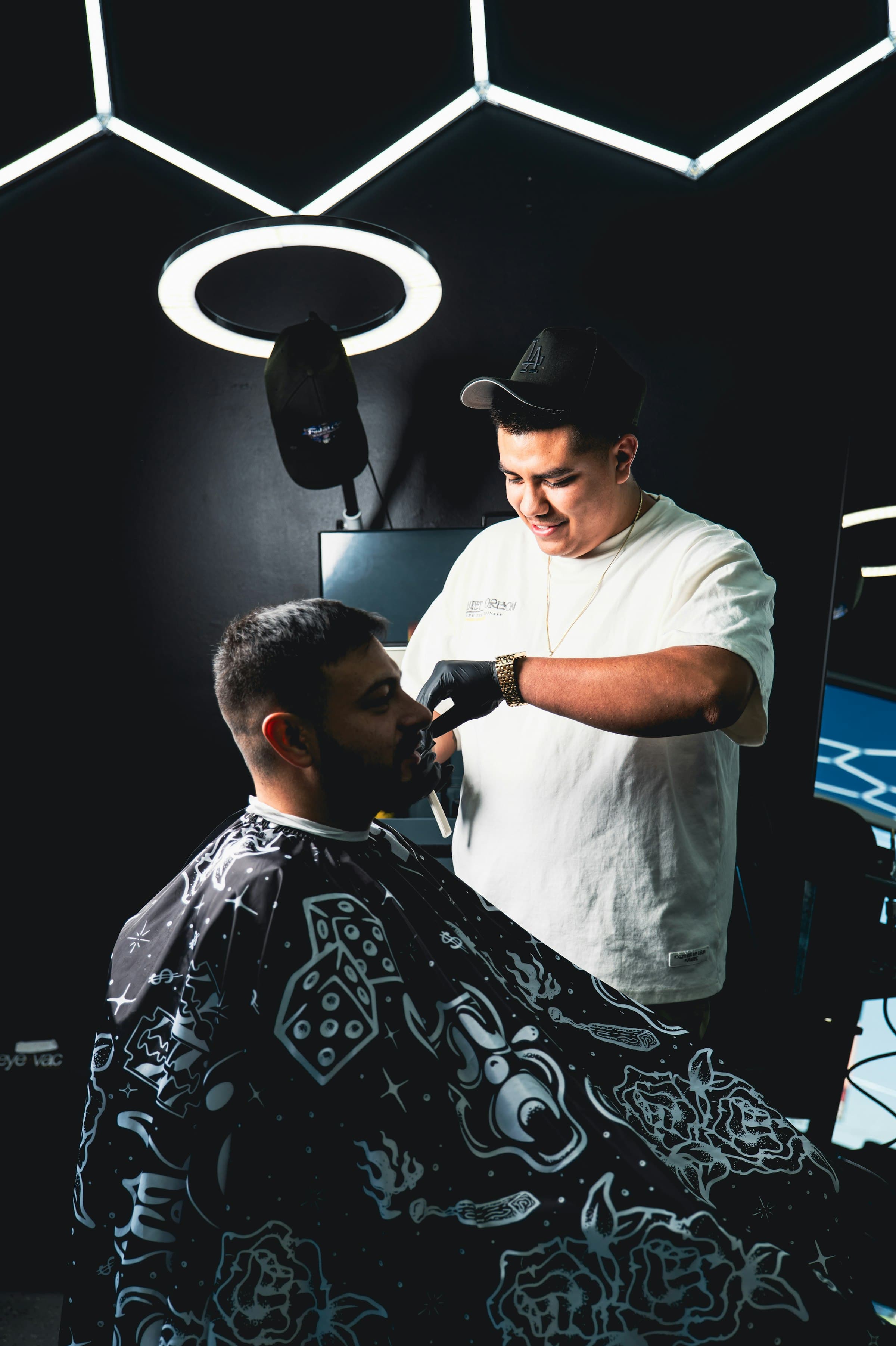 A barber smiling as he trims a client's beard in a dark, modern barbershop